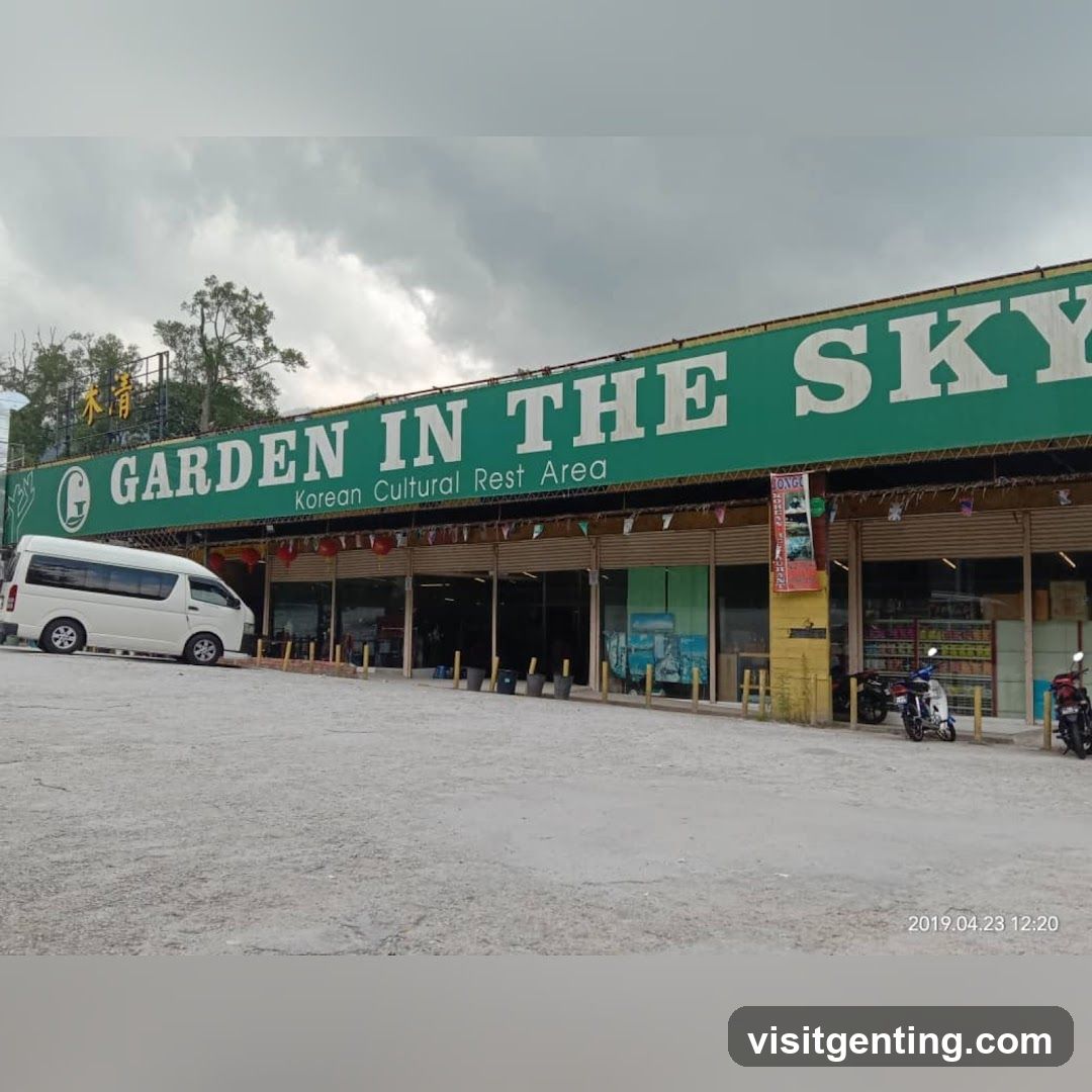 Exterior view of a long green sign with white lettering reading 'GARDEN IN THE SKY' and 'Korean Cultural Rest Area'. The building has a covered entrance area with yellow pillars, glass windows, and red lanterns hanging above. A white van is parked on the left, and several motorcycles are parked on the right. The ground appears to be gravel or dirt. Overcast sky visible in background.