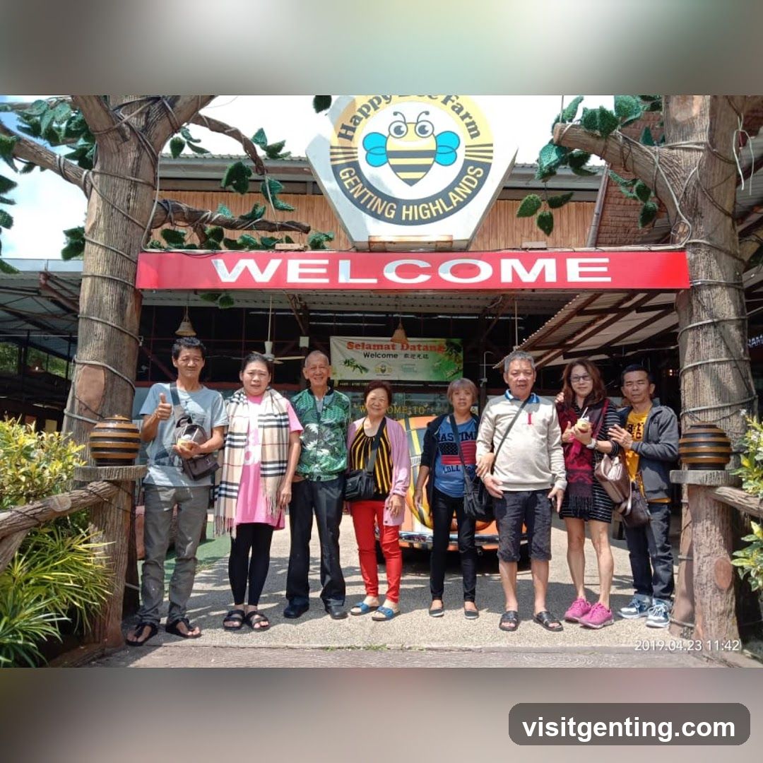 Group of visitors posing in front of a wooden archway entrance with a large red "WELCOME" sign above it. A circular logo featuring a cartoon bee and the text "Happy Bee Farm Genting Highlands" is mounted on the arch. The structure resembles a tree trunk design with decorative elements like hanging pots and artificial leaves. Behind the group, a green banner reads "Selamat Datang Welcome".