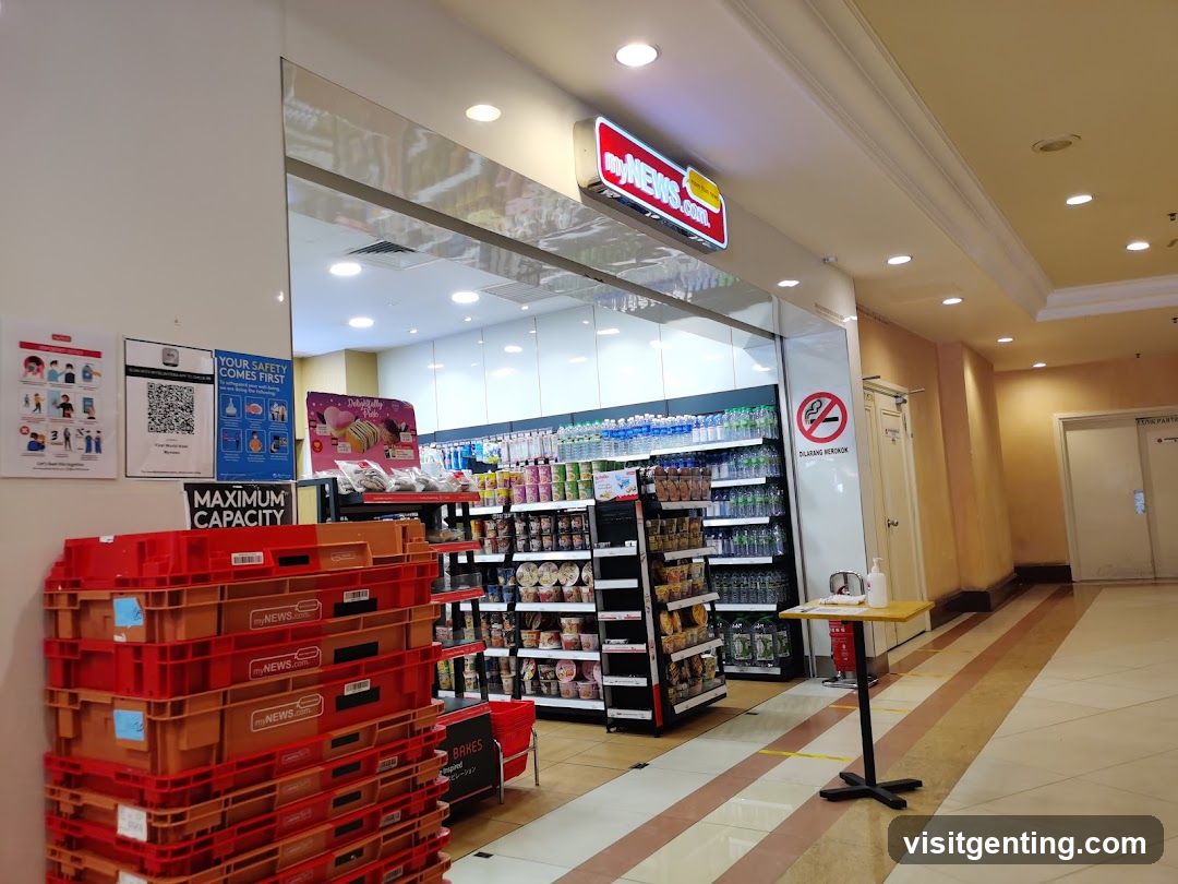 The exterior of a MyNews.com convenience store. The entrance is visible with shelves stocked with drinks and snacks. A prominent red and yellow sign with "myNEWS.com" is displayed above the entrance. To the left, there are posters about safety and maximum capacity, along with stacks of red crates. To the right, a no-smoking sign and a small table with hand sanitizer are present.