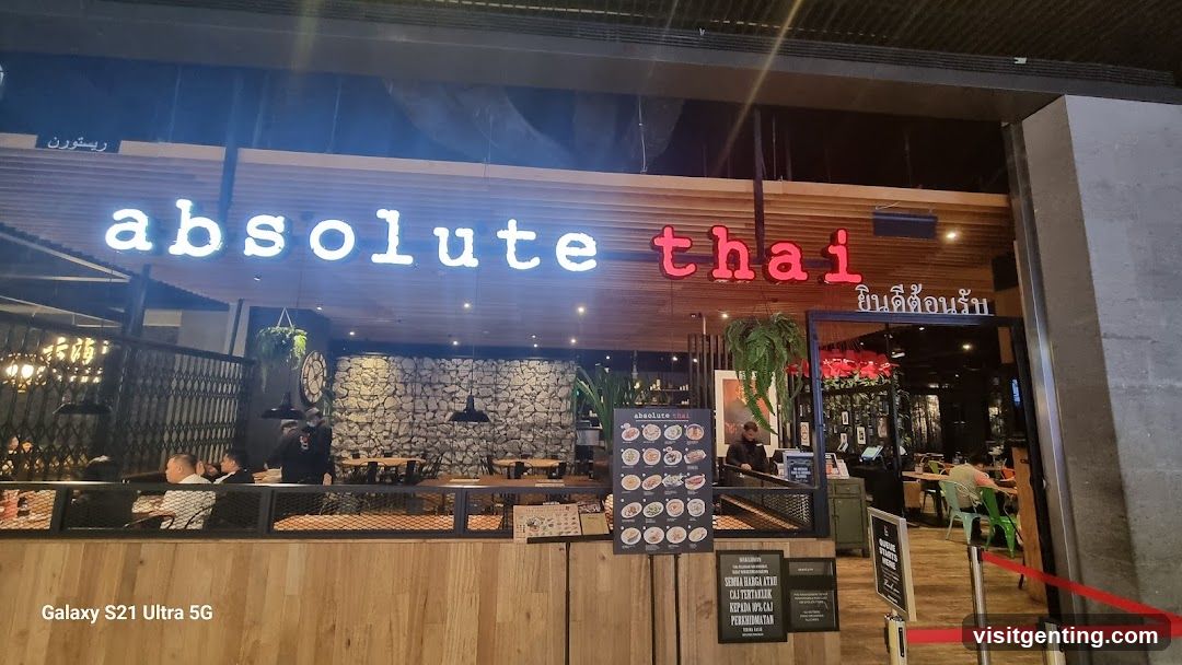 Exterior storefront at night with illuminated sign reading 'absolute thai' in white and red letters. The facade features wood paneling, stone wall accents, hanging plants, and a glass door entrance. Customers are seated inside; staff are visible near the counter. Red velvet rope barriers mark the entryway.
