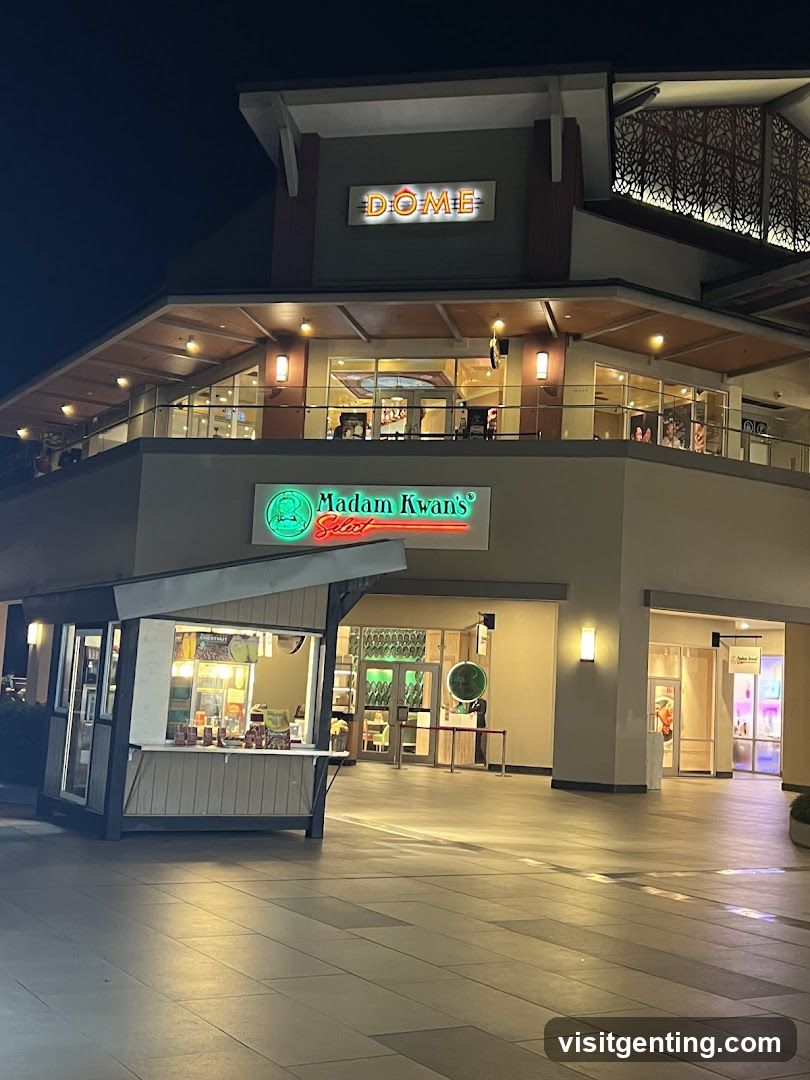 Nighttime exterior view of a multi-level shopping center with illuminated signage. The top level displays 'DOME' in bright letters. Below it, 'Madam Kwan's Select' is lit in green and red neon on the ground floor facade. A small kiosk-style shop is visible to the left under an awning. The entrance area has glass doors and windows showing interior lighting. The pavement is tiled and empty, with no people or staff visible.