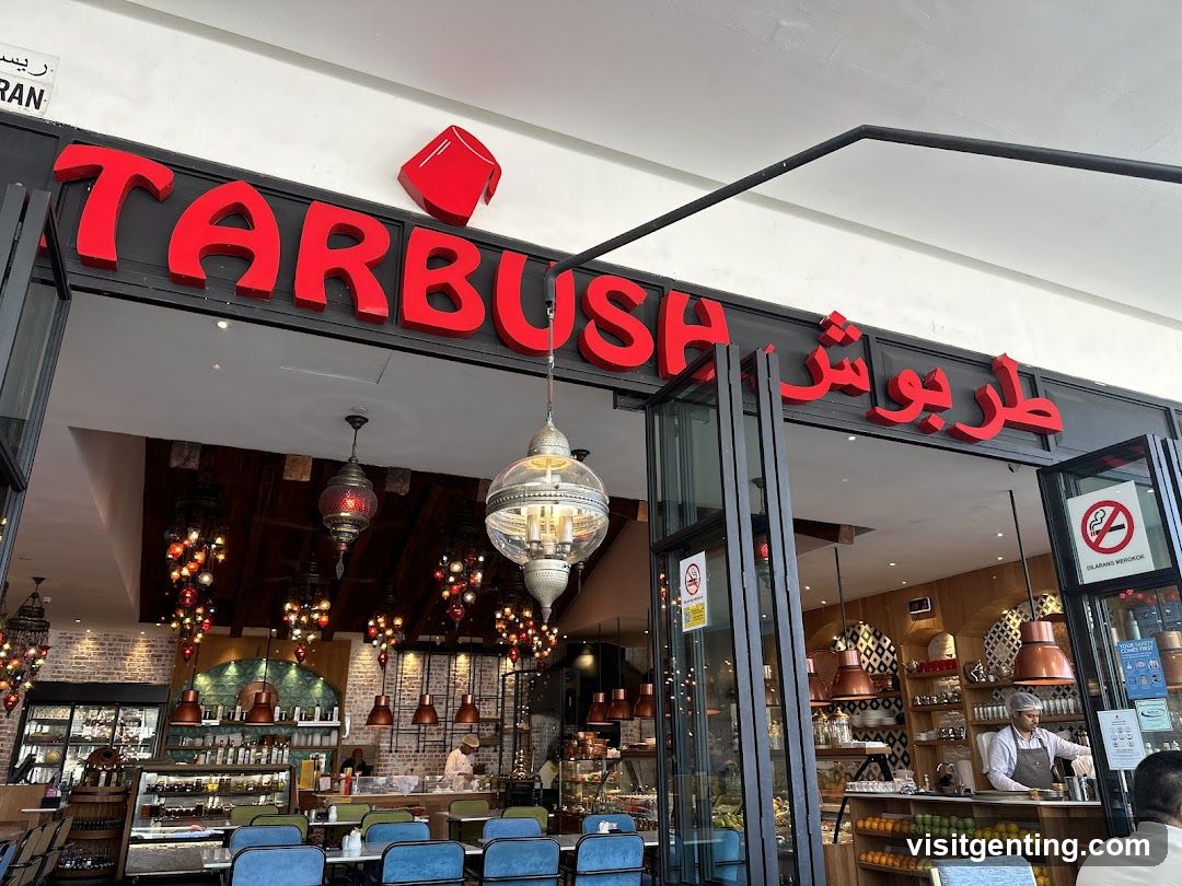 Exterior view of a modern restaurant storefront with prominent red 'TARBUSH' signage in English and Arabic script above the entrance. A stylized red coffee cup icon is mounted on the sign. Glass doors frame the interior, which features warm lighting, hanging lanterns, exposed brick walls, and wooden accents. Staff are visible inside behind counter areas. A no-smoking sign is affixed to the glass door.